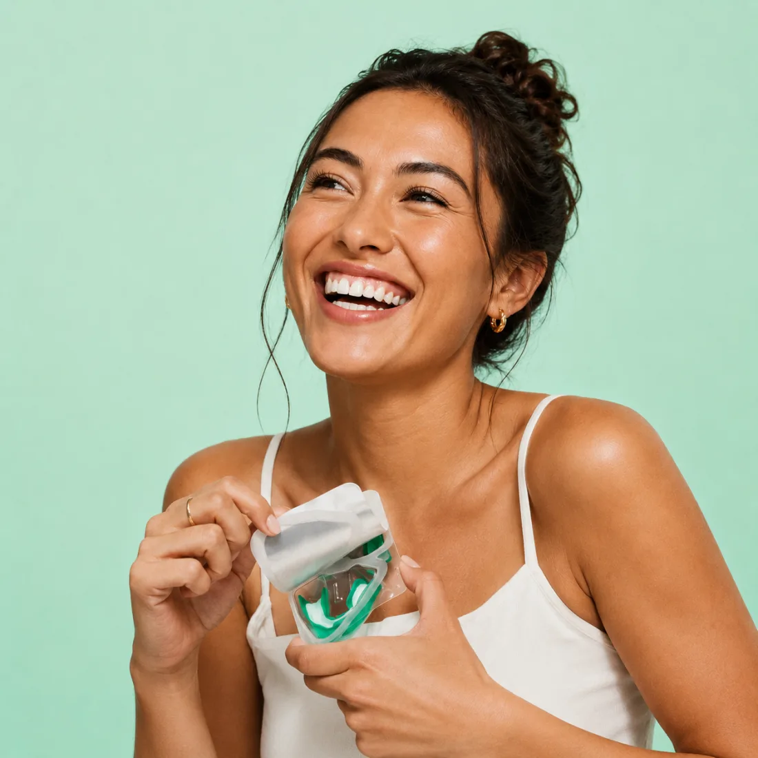 Laughing woman in a white camisole peeling open a clear pack holding a green whitening tray, against a mint backdrop